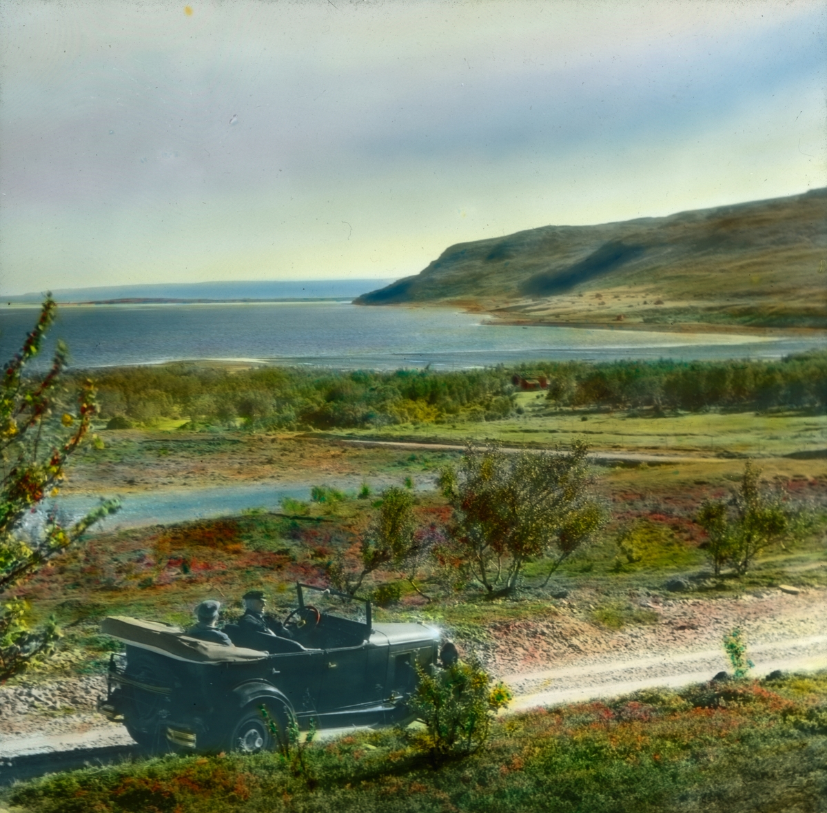 Håndkolorert dias. En åpenbil med en sjåfør og en mann sittende i baksete står på en vei ved Porsangerfjorden.