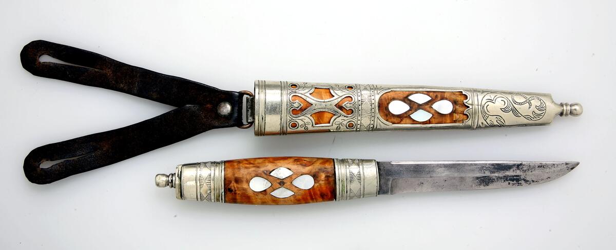 Kniv av Johan Hedenberg med sirlige graveringer, drager i doppsko, bladdekor og perlemor. Skaft og slire i bjørk og nysølv.