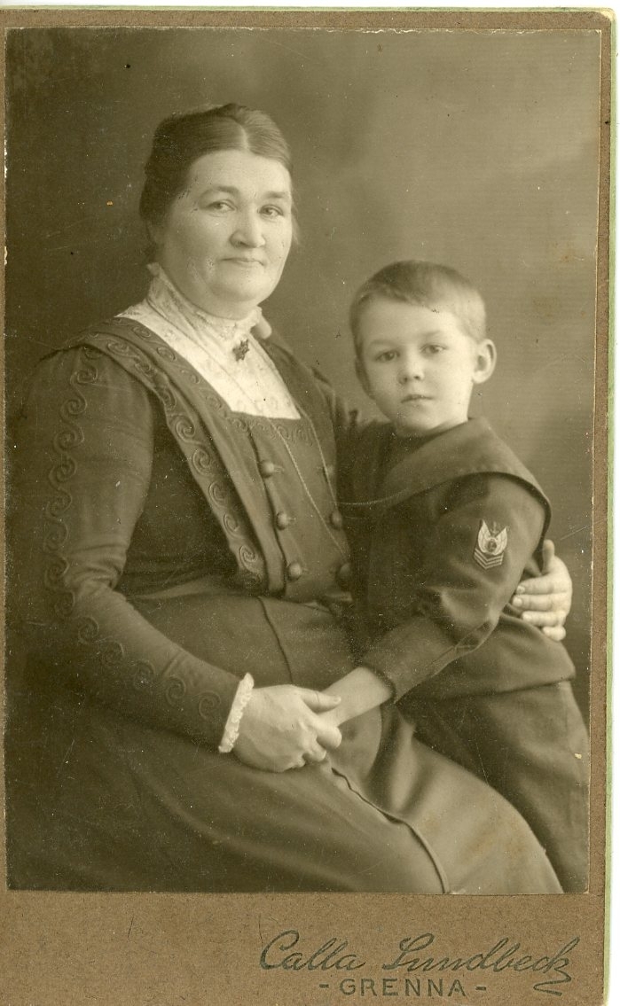 Kabinettsfotografi av en äldre sittande kvinna. Tätt intill henne står en liten pojke, hon håller hans vänstra hand och andra armen om hans midja.