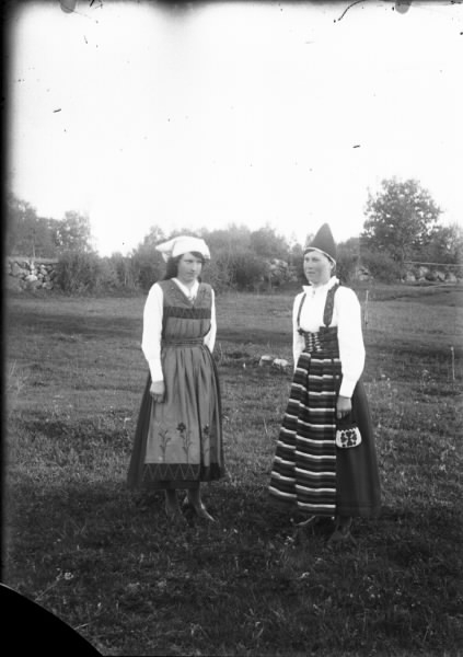 Två unga kvinnor står ute på ett gärde iklädda två olika folkdräkter. (Se även bildnr HB191)