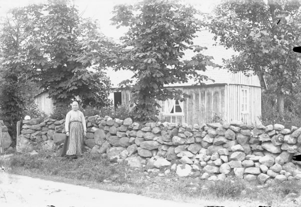 En äldre kvinna står vid en hög stengärdsgård längs vägen som går utanför bostadhuset. I trädgården står kastanjer vid muren.