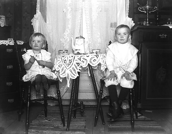 Två barn sitter vid ett litet bord där kanna och koppar dukats fram. Bakom dem hänger spetsgardiner framför en dörr.