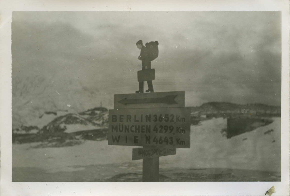 På bildets bakside står det håndskrevet "Wegweiser in Hammerfest", veiviser i Hammerfest. Fotografiet viser en veiviser i vinterlandskap. Skiltene er festet på en firkantet stolpe. Øverst er det festet en utskåren figur av en soldat med stor ryggsekk og koffert. Under den peker en pil til venstre. Det største skiltet informerer om avstandene til Berlin, München og Wien. Nederst viser et skilt veien til "Nordpol 10 km".