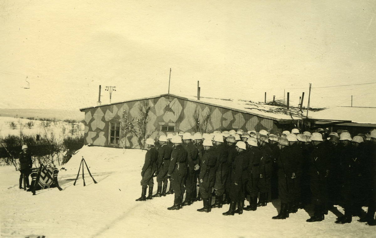 Tyske soldater har tatt oppstilling ved siden av et brakkebygg. Foran dem  står det en soldat bak en hakekorsflagg og gevær som er satt opp tre og tre. Det er vinter. Brakka er mal med et kamuflerende mønster.