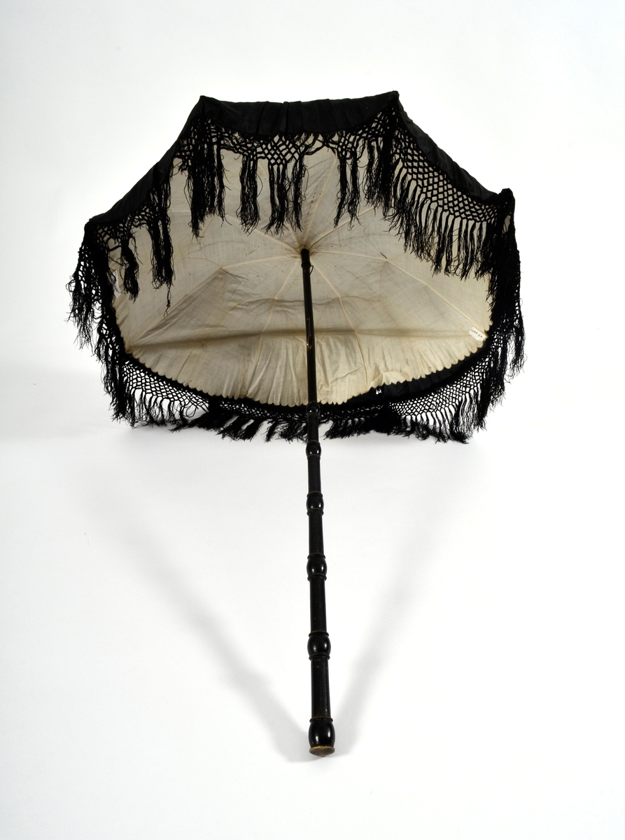 Parasoll i svart silke med frynserbord langs nedrekanten. Fóra med kvitt silketøy. 8 spiler. Stanga er utskåre i svart tre.