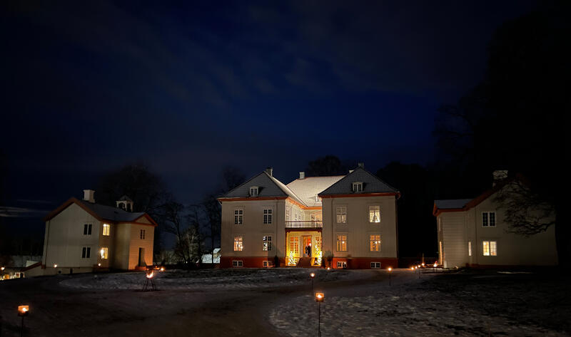 Eidsvollsbygningens hovedfasade og sidebygningene med lys i vindu og på fasaden en mørk vinterkveld