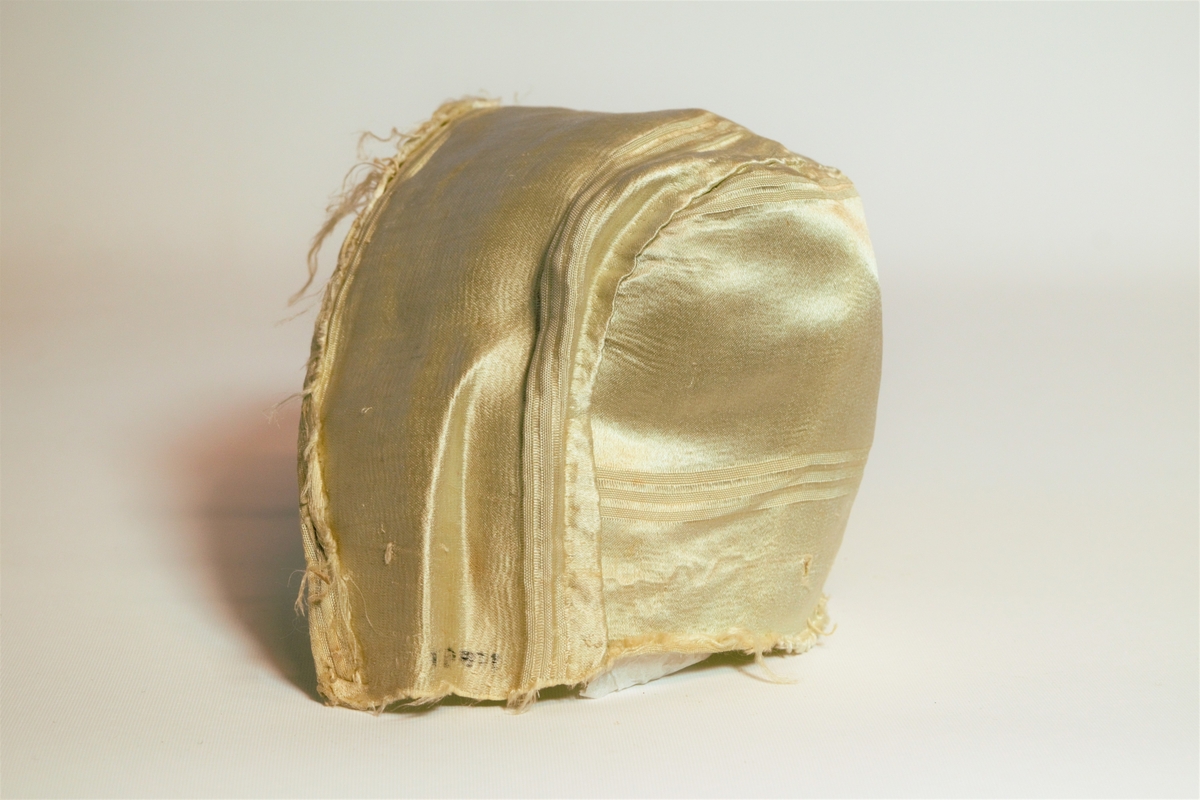 Lue av lys gylden silke, sydd av to tekstilstykker. Begge stykkene har innvevde riller i stoffet. Lua er ikke kantet nederst, den har trolig vært festet til et plagg eller hatt en kappe e. l.