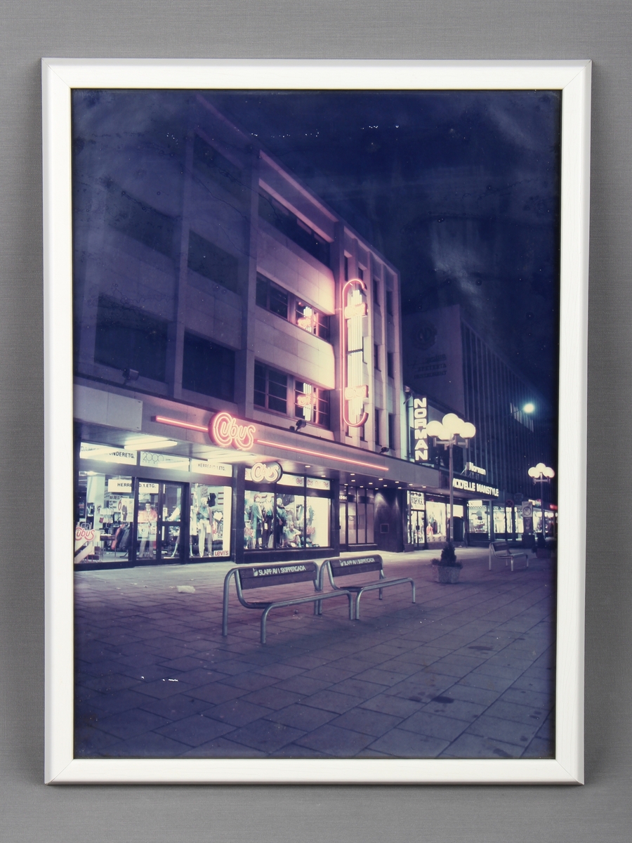 Reklameplakat i ramme, som viser fasaden til en Cubus-butikk om natten.