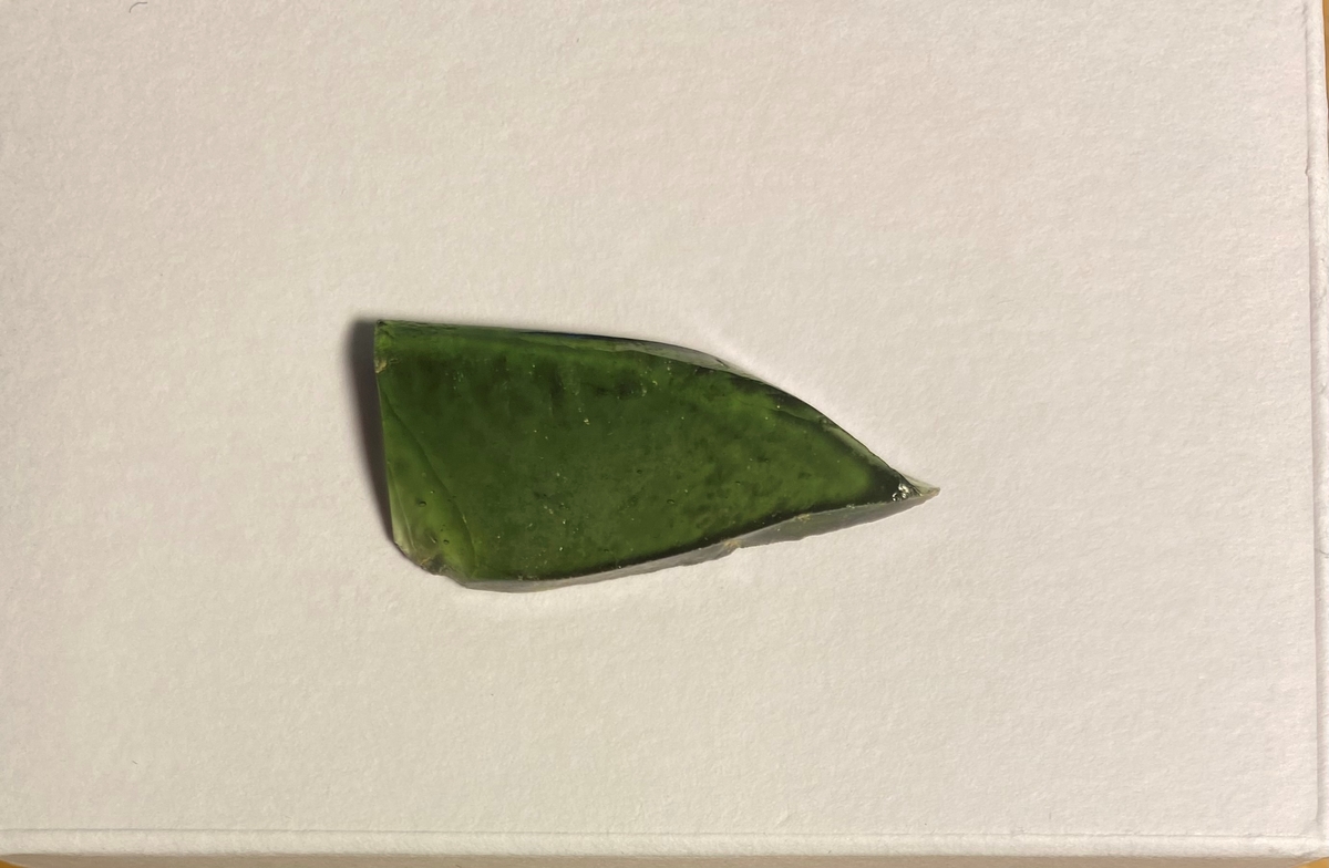 Fragment av ett fönsterglas med mörkgrön massa och små blåsor i glaset. En mjukt rundad kant.