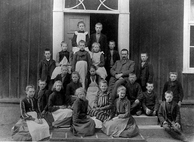 Skolbarn, flickor och pojkar, utanför sin skola i Uppgränna. Den manliga läraren sitter till höger.