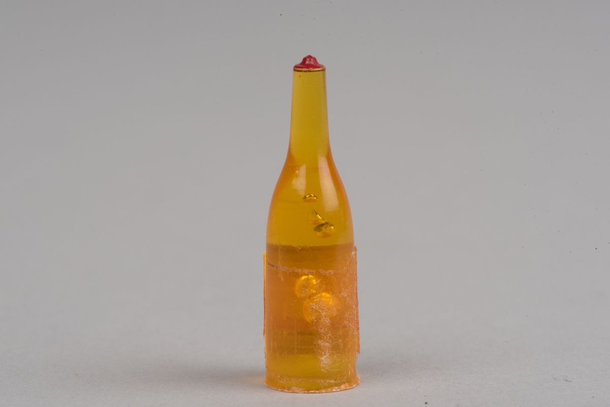 Dockskåpsinredning i form av en whiskyflaska i plast med pappersetikett.
Flaskan är gul med en röd kork. På etiketten framgår att det ska vara en whiskyflaska.