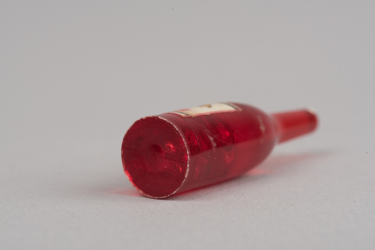 Dockskåpsinredning i form av en vinflaska av plast med etikett.
Flaskan är röd med en vit kork. På etiketten framkommer det att det ska vara en rödvinflaska från Gandesa i Spanien. Etiketten har text och en vindruvsklase.