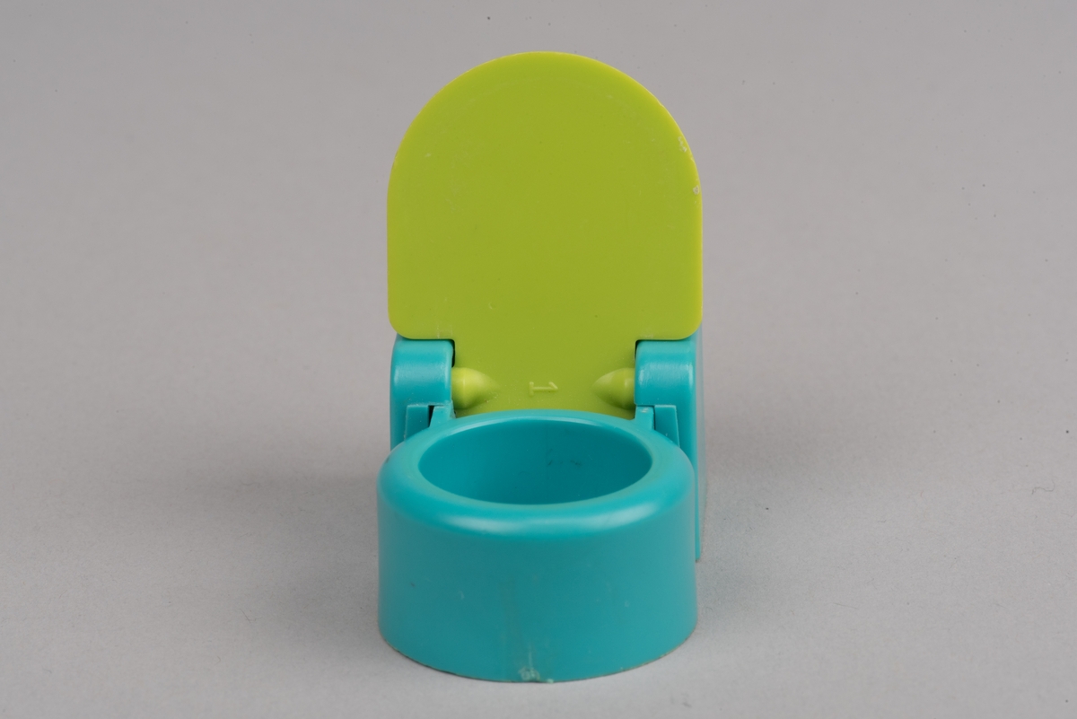 Dockskåpsinredning i form av en toalettstol av plast. Toalettlocket går att fälla upp.
Vattenbehållaren och stolen är turkosa. På ovansidan av behållaren finns en förhöjd spolknapp. Toalettlocket är gulgrönt.