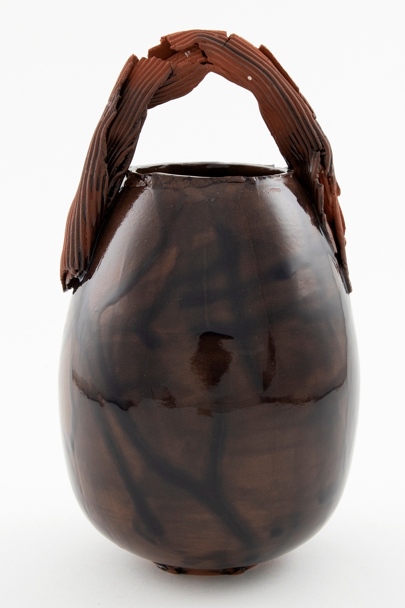 Blank brunglasert vase i terrakotta med eggformet korpus. Lav fot og åpen munning. Vasen har en skulpturell hank i matt brun terrakotta med riller. Hanken går i en bue over munningen.