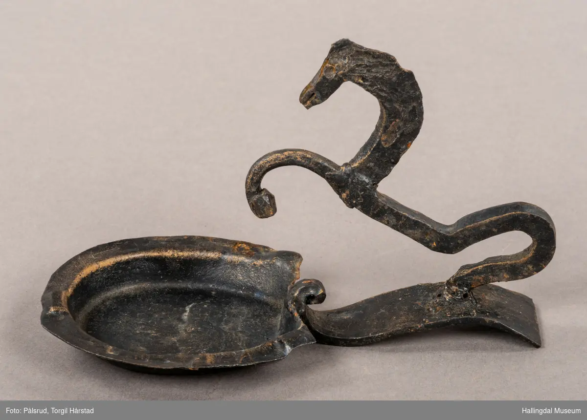 Et askebeger med håndtak i form av en steilende, stilisert hest. Hesteskoformet askebeger. Laget i smijern, og er dekorert med sort og påpusset gull. 