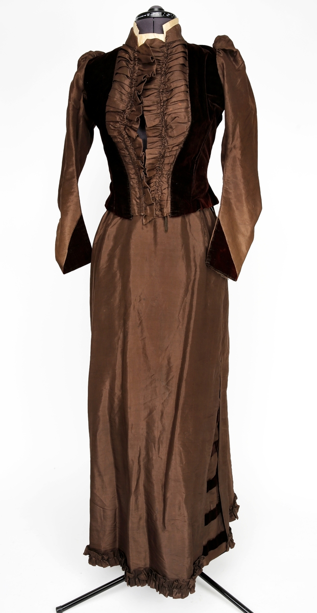 Todelt kjole i silke og fløyel. 
Skjørtet er dekorert med rysjer nederst, splitter med fløyelsdetaljer i sidene, og foldet fløyelsstykke bak med padding ca 25 cm ned fra toppen. 
