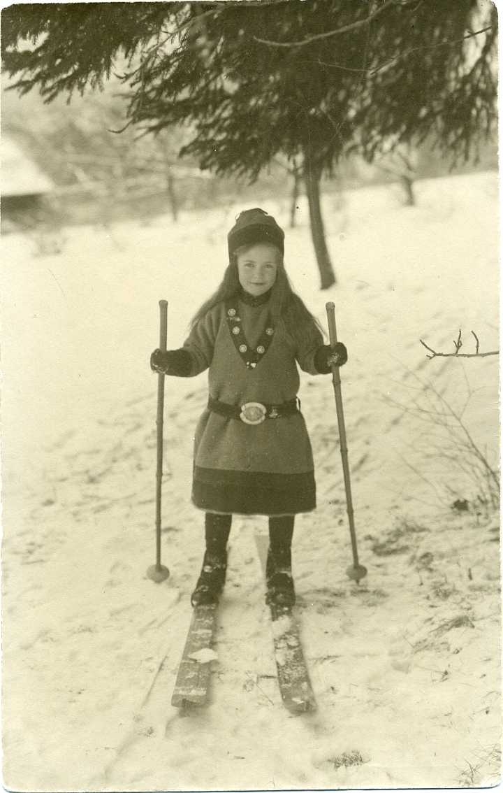 En liten flicka står på en par skidor. Hon bär en mössa och särk i fornnordisk stil. Ett vykort.