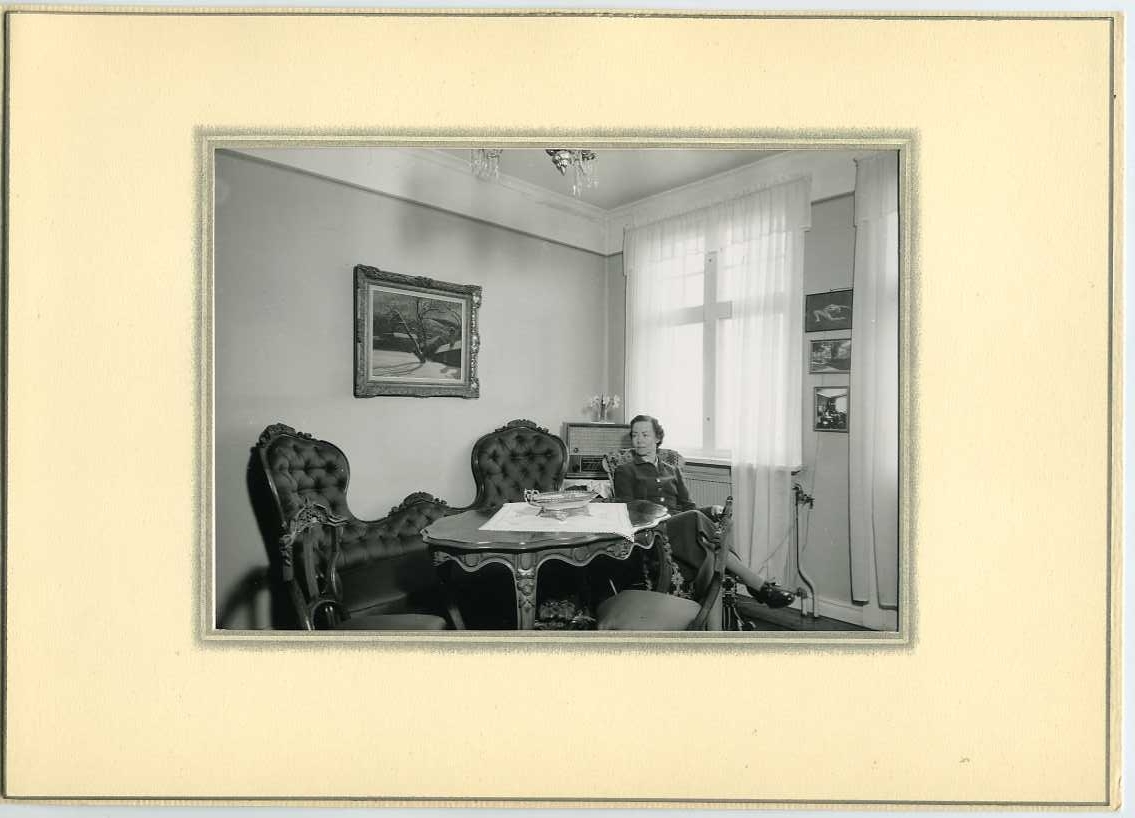 Astrid Malmborg sitter i en fåtölj vid en soffgrupp intill fönster med ljusa, tunna gardiner. Det är i hennes föräldrars hem. På en smal vägg mellan fönsterna sitter tre ramade fotografier/bilder, bland annat en danspose med henne själv. På väggen ovan soffan hänger en målning med grännamotiv av hennes pappa, konstnären Pelle Malmborg.