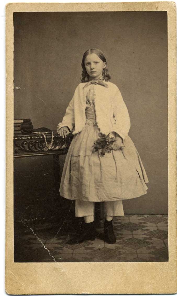 Porträtt av en flicka. Hon står vid ett bord där det ligger böcker och en liten handväska. I vänster hand håller hon en blomsterbukett.