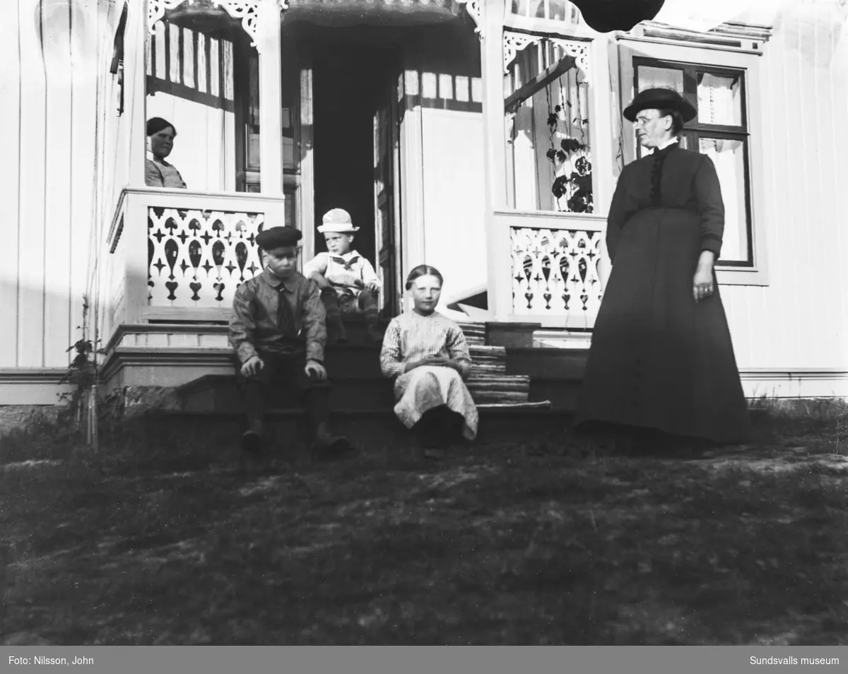 Två bilder med två kvinnor och tre barn på en brokvist med snickarglädje. Huset är ljust målat. Båda bilderna är skadade i överkanten.