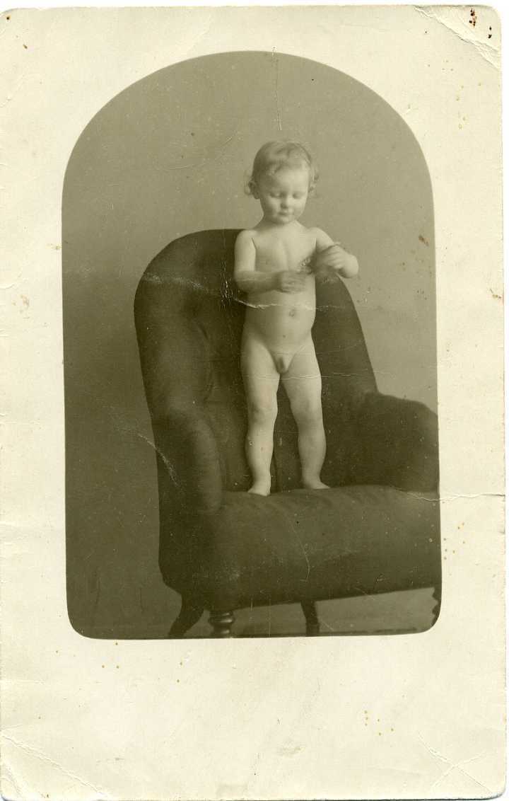 Porträtt av en naken liten pojke som står i en fåtölj. Han håller möjligen en leksak i händerna. Vykort.