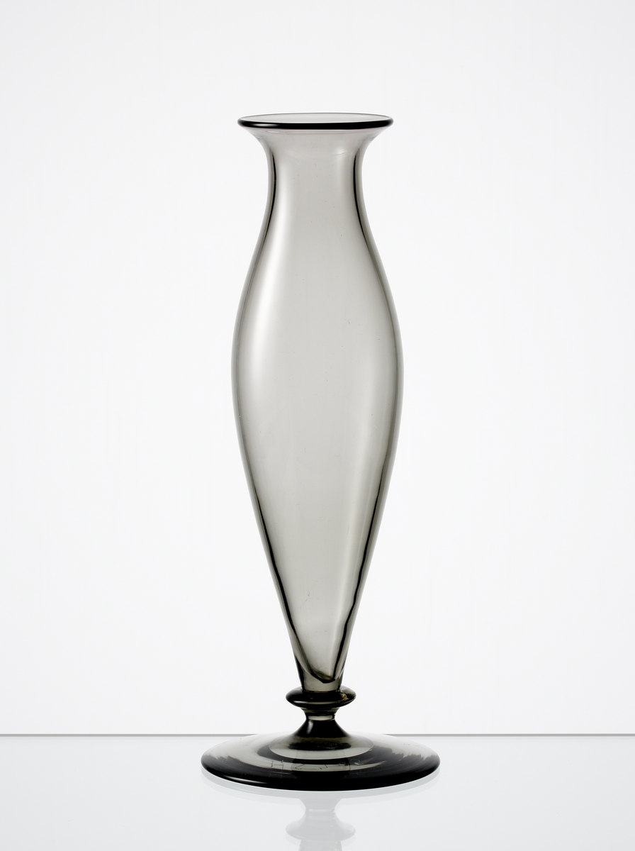 Formgiven av Edward Hald. Blomglas i gråtonat glas. Utsvängd mynning, spolformad kropp med knapp vid foten.