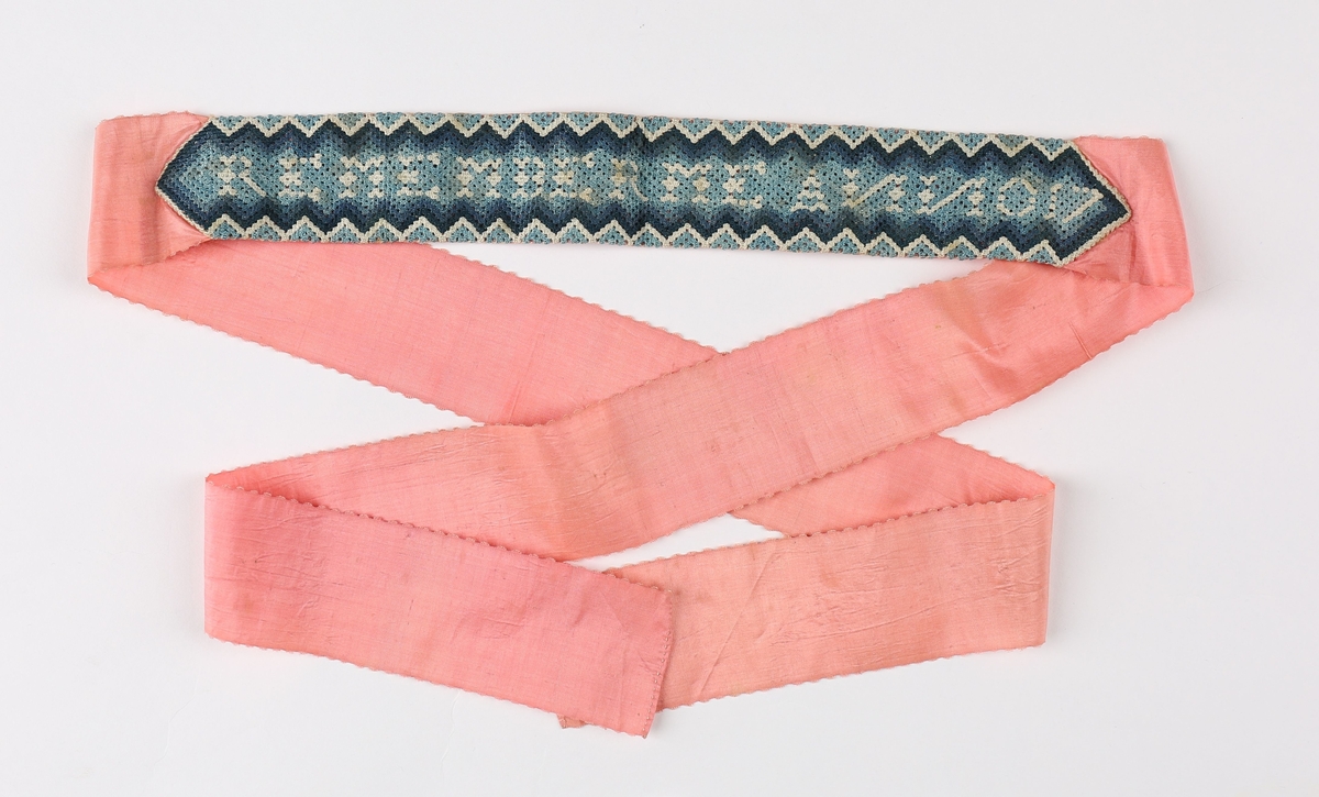 Av rosa silkerips uthakket langs kanten og smal fall for endene. På midten er påsydd en bord av fin, tett hekling i tre blåfarger og hvitt. Mønsteret består av langsgående sikk-sakkborder og en innskrift på midten.