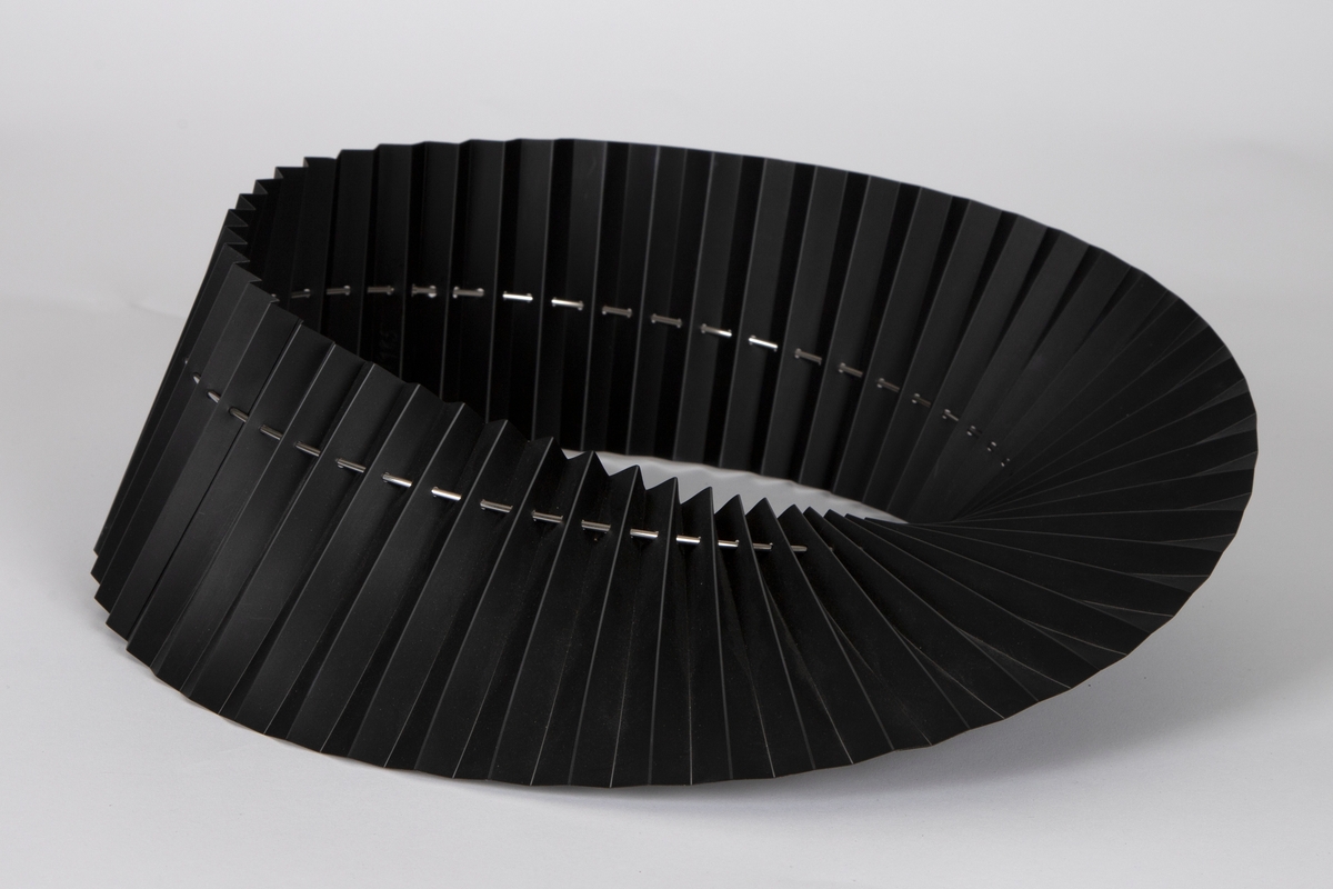 Halskrage av foldelagt svart PVC som er tredd på en sirkelrund stålring og er noe vridd i forhold til denne.