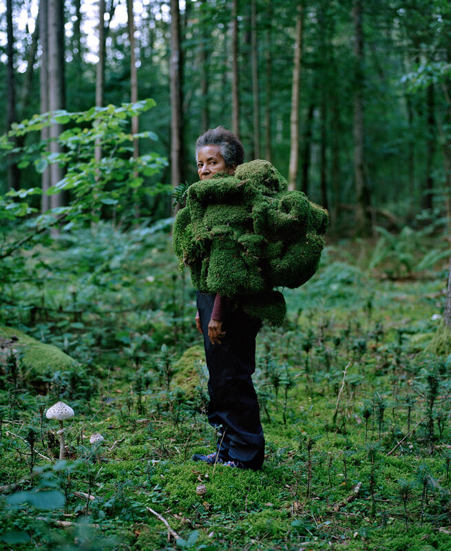 En kvinne står midt i en skog. Hun er delvis kledd i en drakt av mose. Hun ser rett på oss. Fotografi.