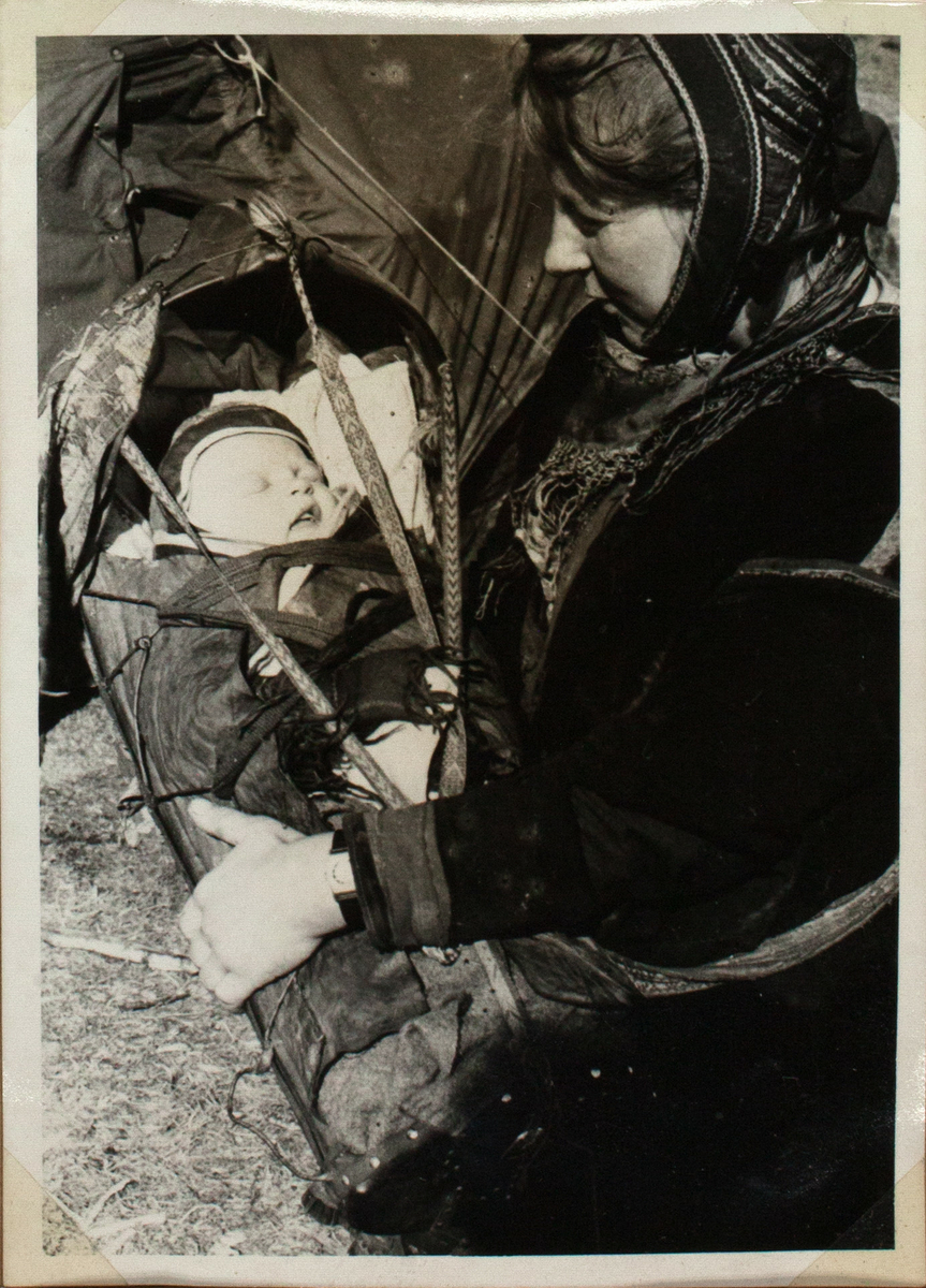 Bildet viser en samisk kvinne som holder en komse med spedbarnet inn i. Babyen sover men hans mor ser ned på den. Kvinne sitter ved siden av et telt og er ikledd tradisjonelle samiske antrekk.