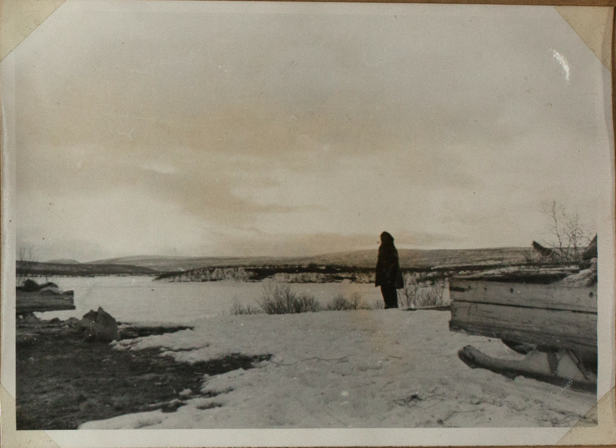 Bildet viser en person som men står se over et snødekket vann. Landskapet er delvis snødekket. I forgrunnen til høyre står det en slede.