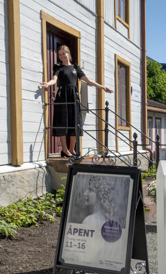 Ung kvinne i svart kjole står på trappa til Kirsten Flagstad museum og slår inviterende ut med armene for å ønske velkommen. Nedenfor trappa står en plakatbukk med en plakat med bilde av en ung Kirsten Flagstad.