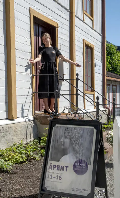 Ung kvinne i svart kjole står på trappa til Kirsten Flagstad museum og slår inviterende ut med armene for å ønske velkommen. Nedenfor trappa står en plakatbukk med en plakat med bilde av en ung Kirsten Flagstad.