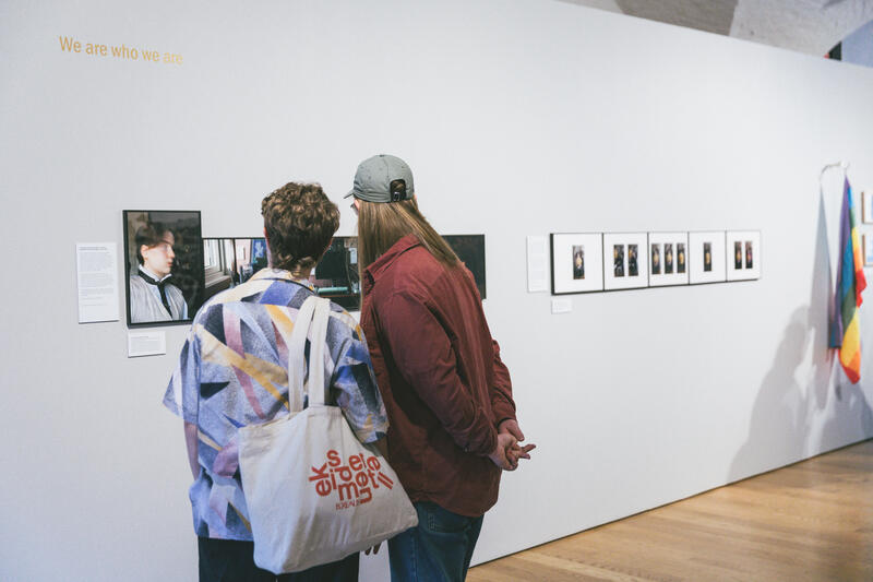 To personer, sett bakfra, ser på bilder på en utstillingsvegg. Fotografi.