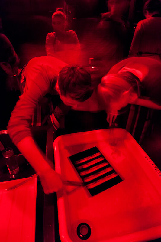 Bildet er rødt og svart og viser personer som jobber i et mørkerom. De er konsentrert over et kar der det står skrevet "fremkaller" på kanten. Rommet er mørkt og lyset er rødt. Fotografi.