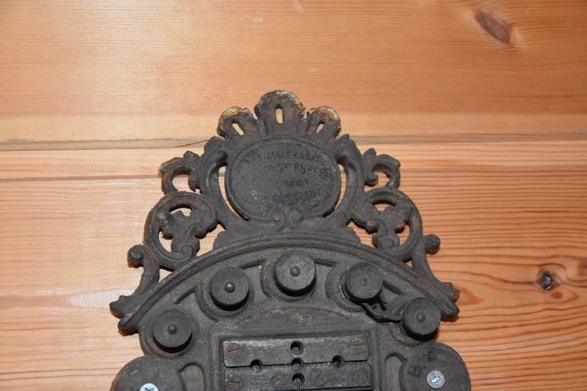 Magneto veggtelefon av blikk med ornamentert støpejern. Treimitasjon og Geometriske dekorasjoner på lokk og fremside som kan minne om treimitasjon. Stoffledning som er tvunnet. Tekst i øvre del. Sveiv på høyre side. Røret med høytaler og"taltetut" henger på venstre side.