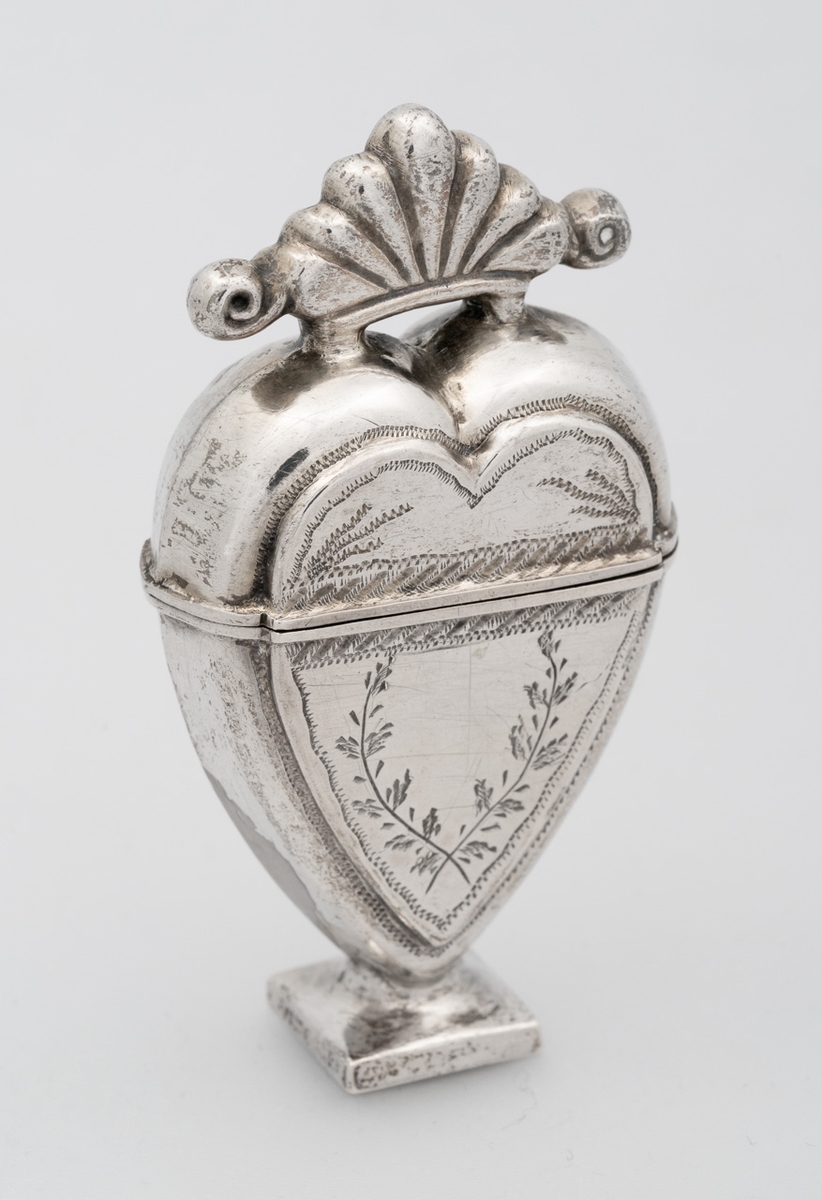 Hjerteformet luktevannshus i sølv med firkantet fot og et skjellornament med volutter på lokket. Korpus er dekorert med kryssende grener på forsiden, samt medaljong på baksiden. Innvendig forgylt.