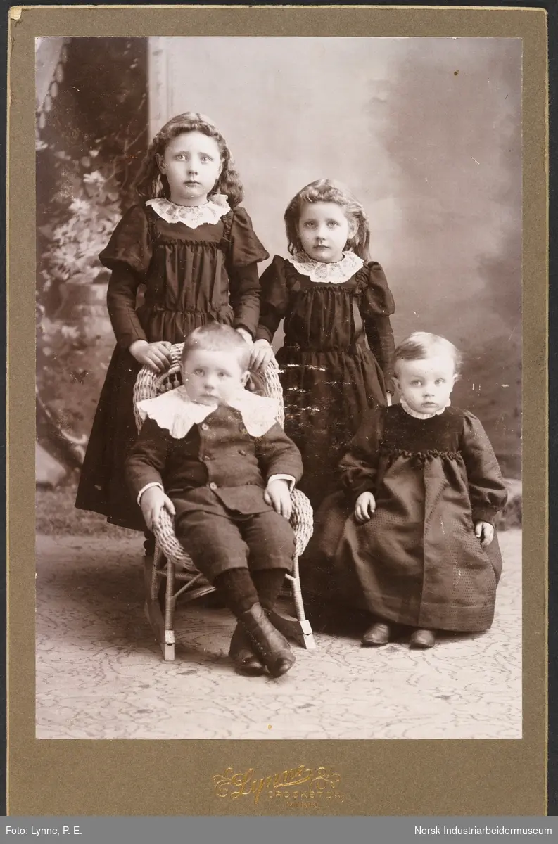 Visittkortfoto, fire barn fotografert i studio. Tre jenter kledd i kjoler og en gutt i matrosdress sittende i kurvstol. Den yngste jenta sitter i stol, de to eldste står bak.