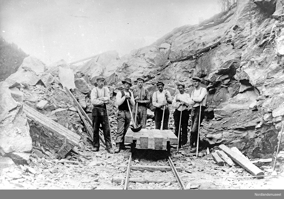 Arbeidere med spett og slegge. Fjellmasse bak dem, jernbaneskinner i forgrunnen. Arbeidsgjeng i Sulitjelma. Fra venstre: 1. ukjent, 2. Didrik Johansen, 3. Kristen Johansen, 6. Kristen Gåre.