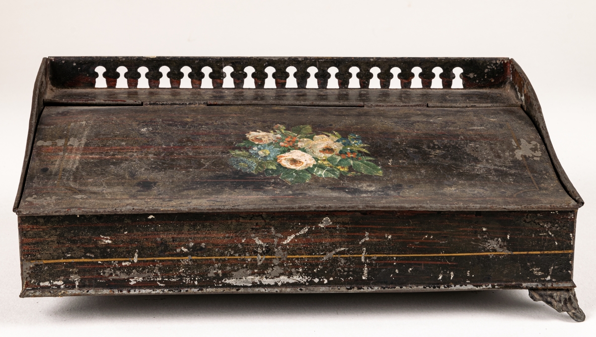 Eventuellt skrivställ, i pulpetform, av plåt. Låda med lutande lock, svart-brunt med blomstermålning.