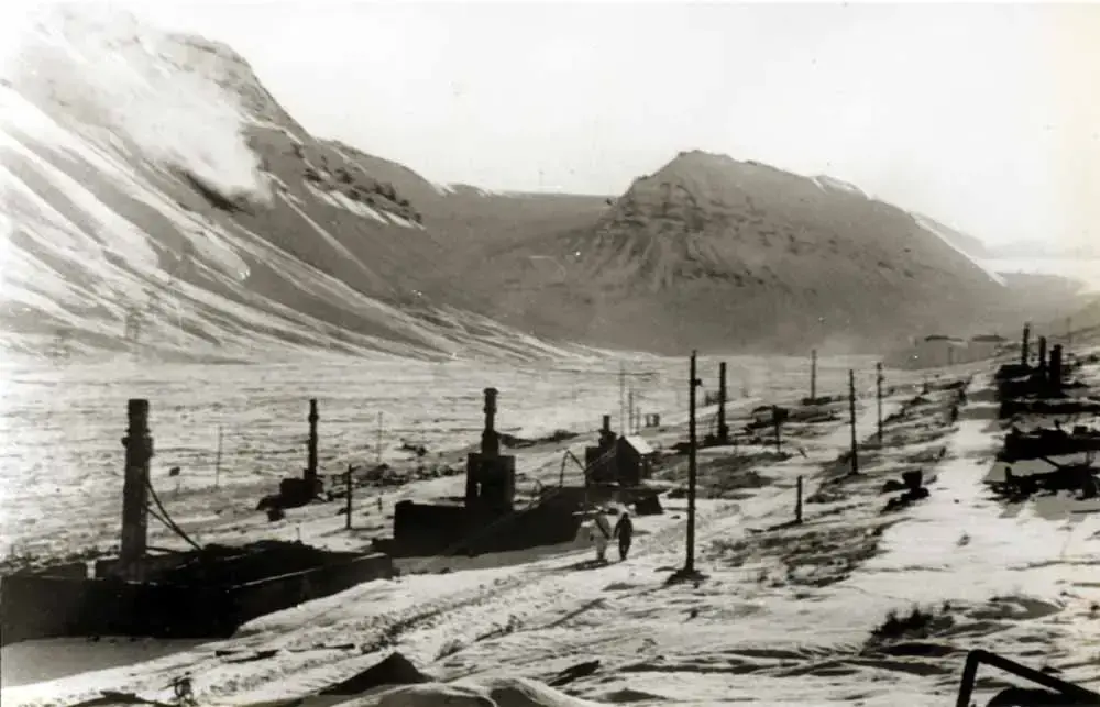 Nedbrente brakker, hvor kun ovn/peis står igjen. Fjell og sol i bakgrunnen.