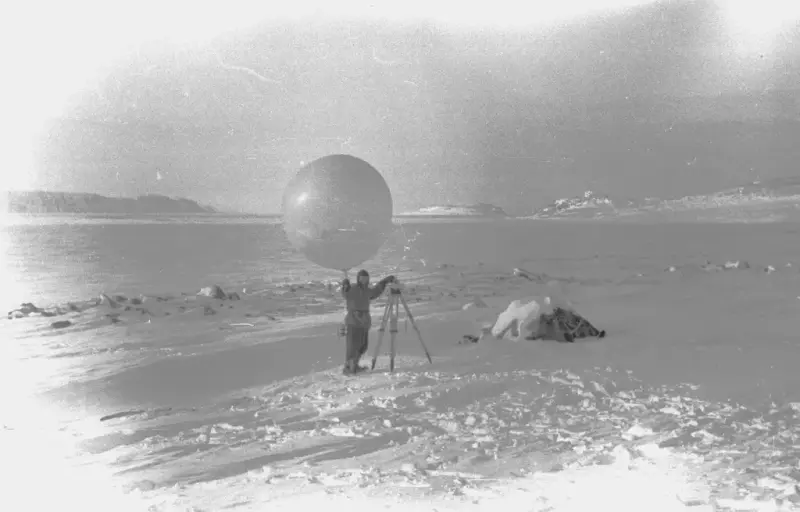 Fotografi av mann som blåser opp en hvit værballong. Ved siden av han står trefot. Landskapet er dekket av snø og is.