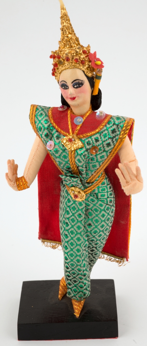 Kvinnefigur i sør-øst-asiatisk glitrende grønn kjole og tårnaktig hodepynt. Står på svart tresokkel.