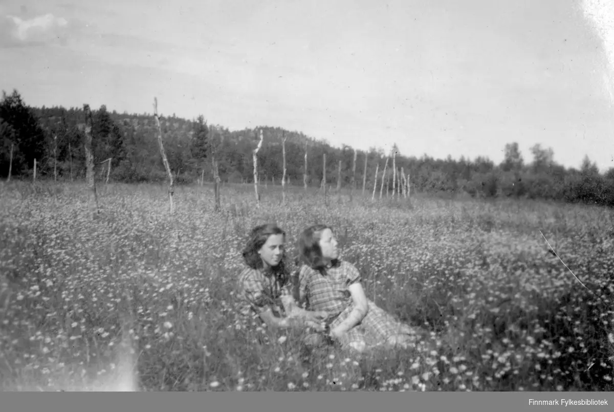 To unge kvinner sitter i blomstereng. Stedet er Løvli i Pasvik, sommeren 1947.