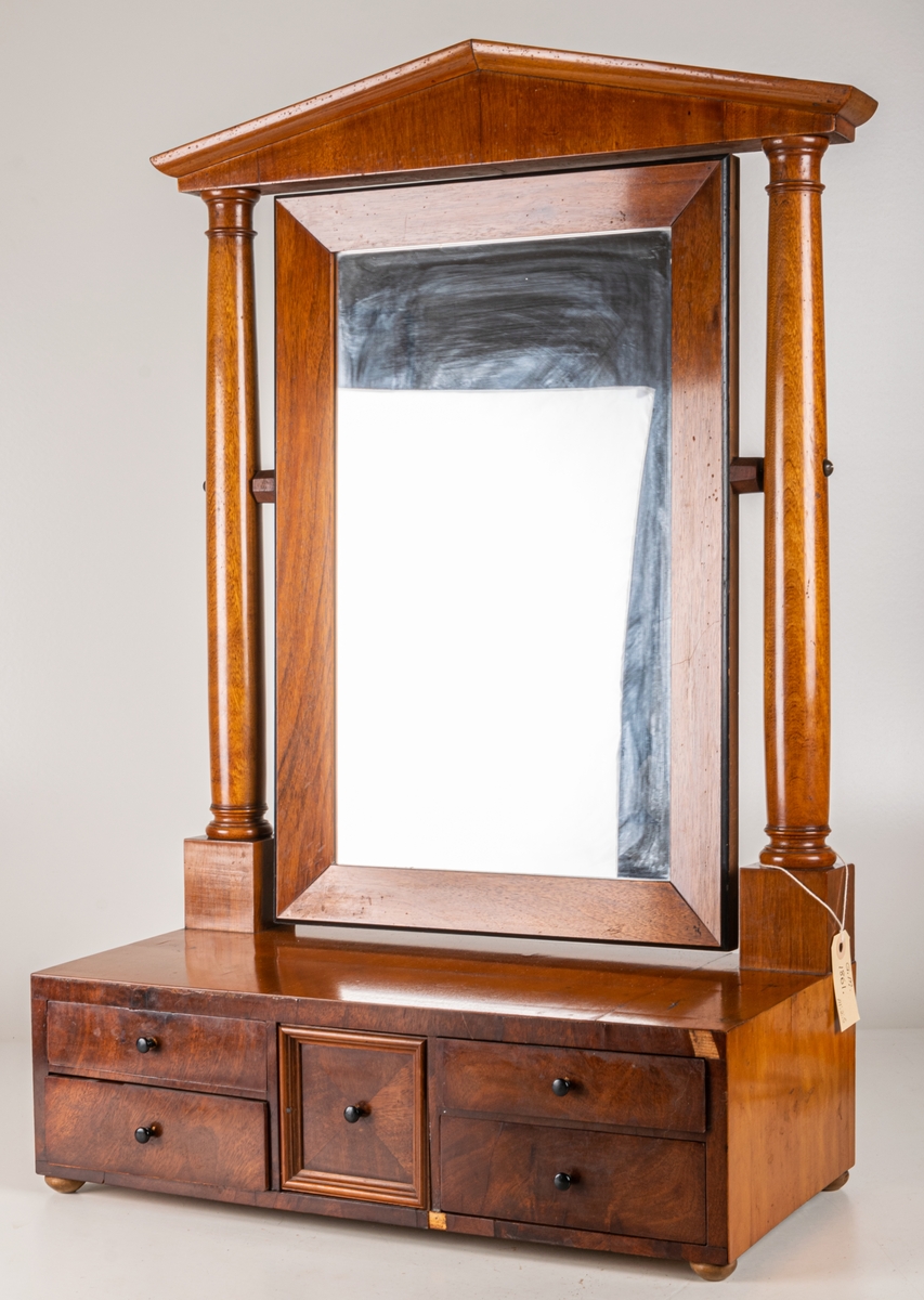 Toilettspegel, Karl-Johanstil, 1800-talet början. Av mahogny, med lådor. Spegeln rörlig mellan två pelare med överstycke. Spegellåda innehållande en större mittlåda och 4 smalare sidolådor.