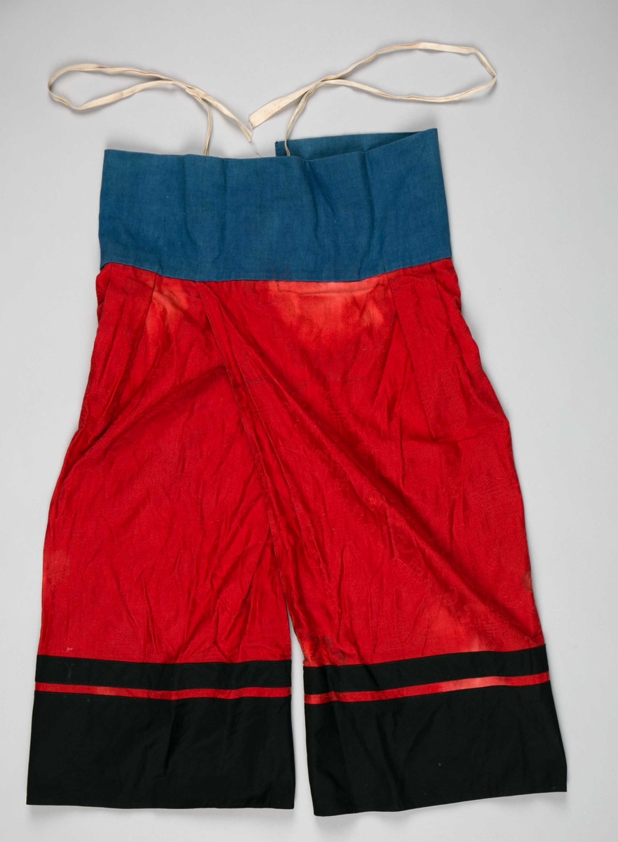 Kinesik knyttebukse i rød silkebrokade til barn ca. 4-5 år. Benpartiet er kantet med svart bomullsstoff. I livet er buksen kantet med himmelblått bomullsstoff.