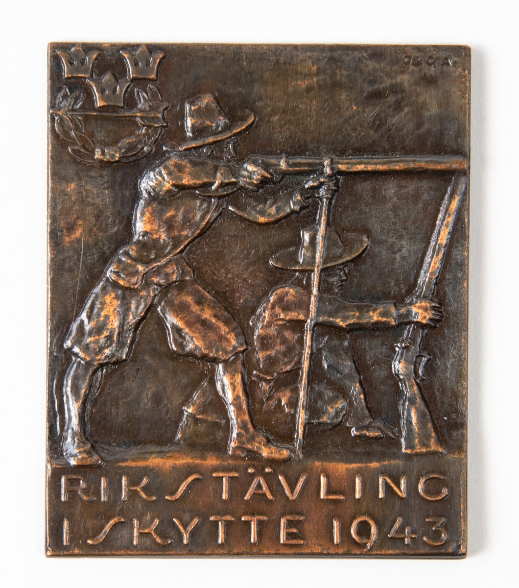 Bronsplakett, Rikstävling i skytte 1943. Motiv med en relief föreställande två män med gevär, en sitter på knä, en står upp och siktar. I övre vänstra hörnet finns en krans med en pil i, samt tre kronor över.