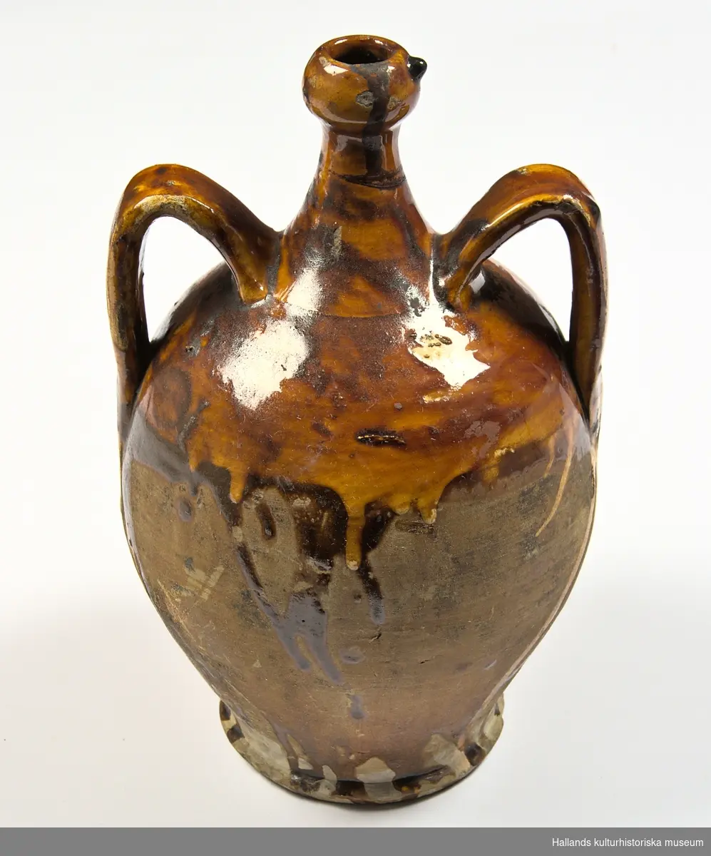 Krus, (urna) av keramik. Har två öron, (handtag) och smal hals. Med glaserad överdel, gulbrun, flytande glasyr.