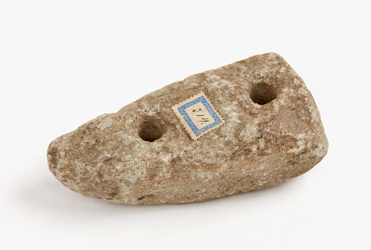 Sten med två plana sidor och två hål borrade genom stenen, 11 mm diam. Sannolikt ej förhistorisk.