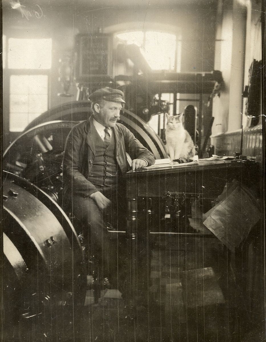 En man i skärmmössa sitter och pratar med en katt vid en pulpet, nära en generator.
Maskinmästare Johan Åström, trol. på elverket i Växjö ca 1920-1925.